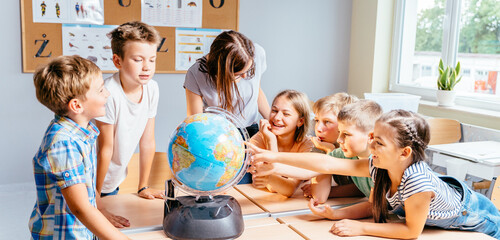 Canvas Print - Happy cute enthusiastically pupils smiling around a globe in classroom with female teacher at school. Group of curious classmates at workplace studying globe in classroom.