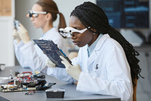Portrait of young black woman wearing magnifying glasses and inspecting electronic parts in quality control lab
