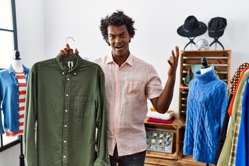 Sticker - African man with curly hair holding shirt from clothing rack at retail shop celebrating victory with happy smile and winner expression with raised hands