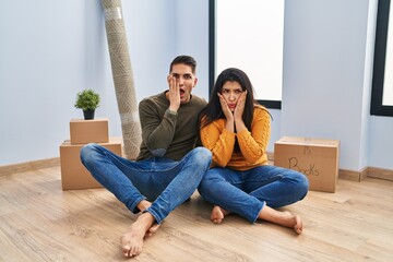 Poster - Young couple sitting on the floor at new home afraid and shocked, surprise and amazed expression with hands on face