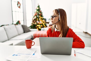 Canvas Print - Young caucasian girl sitting on the table working using laptop by christmas tree looking to side, relax profile pose with natural face with confident smile.