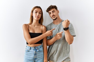 Poster - Young beautiful couple standing together over isolated background in hurry pointing to watch time, impatience, looking at the camera with relaxed expression