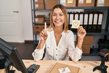 Poster - Young blonde woman working at small business ecommerce holding heart note smiling with an idea or question pointing finger with happy face, number one