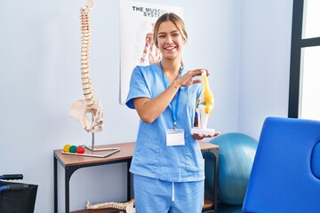 Poster - Young blonde orthopedic woman holding anatomical model of knee joint smiling and laughing hard out loud because funny crazy joke.