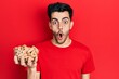 © Krakenimages.com - Young hispanic man holding peanuts scared and amazed with open mouth for surprise, disbelief face