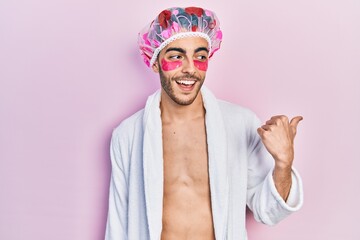 Young hispanic man wearing bathrobe and shower cap using eyes bags patches pointing thumb up to the side smiling happy with open mouth