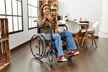 Young Hispanic Girl Sitting On Wheelchair At Home Smelling Something Stinky And Disgusting, Intolerable Smell, Holding Breath With Fingers On Nose. Bad Smell