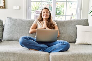 Sticker - Beautiful young brunette woman sitting on the sofa using computer laptop at home praying with hands together asking for forgiveness smiling confident.