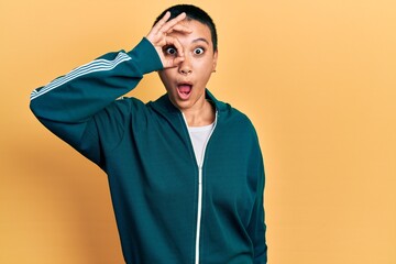 Poster - Beautiful hispanic woman with short hair wearing sporty jacket doing ok gesture shocked with surprised face, eye looking through fingers. unbelieving expression.