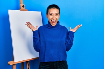 Sticker - Beautiful hispanic woman with short hair standing by painter easel stand celebrating mad and crazy for success with arms raised and closed eyes screaming excited. winner concept