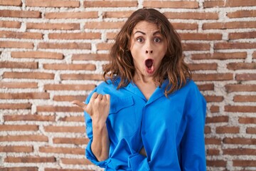 Poster - Beautiful brunette woman standing over bricks wall surprised pointing with hand finger to the side, open mouth amazed expression.