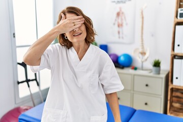 Canvas Print - Middle age physiotherapist woman working at pain recovery clinic smiling and laughing with hand on face covering eyes for surprise. blind concept.