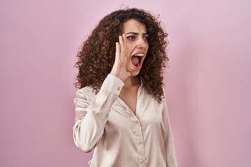 Sticker - Hispanic woman with curly hair standing over pink background shouting and screaming loud to side with hand on mouth. communication concept.