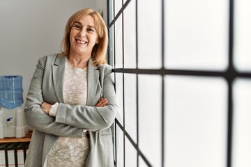 Wall Mural - Middle age businesswoman standing with arms crossed gesture leaning on the window at the office.