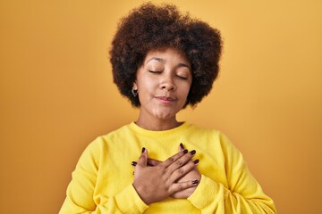 Sticker - Young african american woman standing over yellow background smiling with hands on chest with closed eyes and grateful gesture on face. health concept.