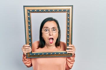 Poster - Middle age hispanic woman holding empty frame afraid and shocked with surprise and amazed expression, fear and excited face.