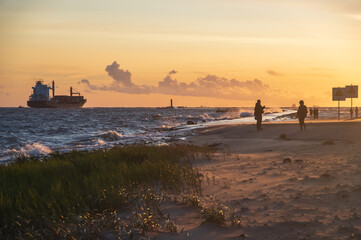 Wall Mural - Soft focus image of ship leaving the harbor on windy sunny evening passing the lighthouse. Silhouettes of people walking by the mole