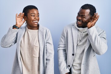 Sticker - Young african american couple standing over blue background together smiling with hand over ear listening an hearing to rumor or gossip. deafness concept.