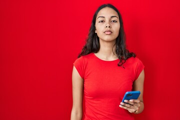 Canvas Print - Young brazilian woman using smartphone over red background relaxed with serious expression on face. simple and natural looking at the camera.