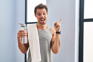 Poster - Young hispanic man wearing sportswear drinking water with a big smile on face, pointing with hand finger to the side looking at the camera.