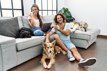 Poster - Young hispanic couple with dogs relaxing at home with a big smile on face, pointing with hand finger to the side looking at the camera.