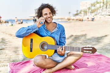 Sticker - Young hispanic man playing guitar and talking on the smartphone sitting on sand at the beach.