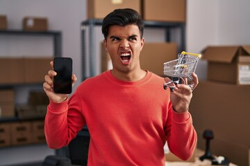 Poster - Hispanic man working at small business ecommerce holding cart angry and mad screaming frustrated and furious, shouting with anger. rage and aggressive concept.