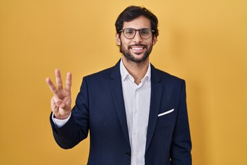 Sticker - Handsome latin man standing over yellow background showing and pointing up with fingers number three while smiling confident and happy.