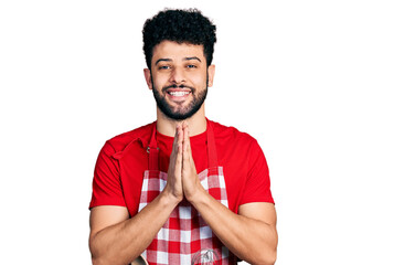 Poster - Young arab man with beard wearing cook apron praying with hands together asking for forgiveness smiling confident.