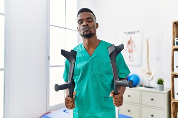 Wall Mural - Young african physiotherapist man holding crutches looking at the camera blowing a kiss being lovely and sexy. love expression.