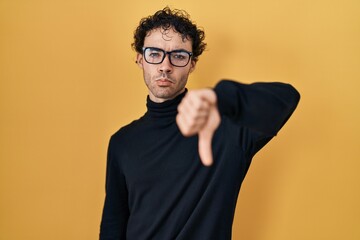 Poster - Hispanic man standing over yellow background looking unhappy and angry showing rejection and negative with thumbs down gesture. bad expression.