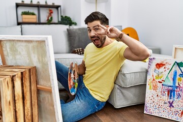 Poster - Young man with beard painting canvas at home doing peace symbol with fingers over face, smiling cheerful showing victory