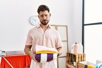 Poster - Young man with beard holding folded laundry after ironing skeptic and nervous, frowning upset because of problem. negative person.