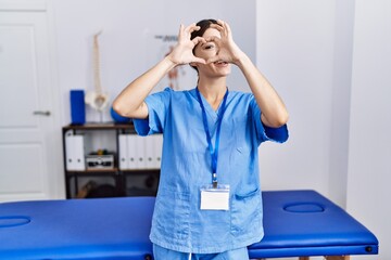 Poster - Young hispanic woman wearing physiotherapist uniform standing at clinic doing heart shape with hand and fingers smiling looking through sign