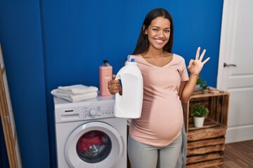 Sticker - Young pregnant woman doing laundry holding detergent bottle doing ok sign with fingers, smiling friendly gesturing excellent symbol