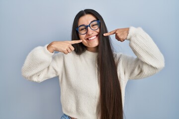 Poster - Young hispanic woman wearing casual sweater over blue background smiling cheerful showing and pointing with fingers teeth and mouth. dental health concept.