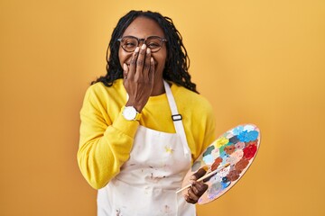 Canvas Print - African woman holding painter palette laughing and embarrassed giggle covering mouth with hands, gossip and scandal concept