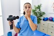 Young physiotherapist woman holding therapy massage gun at wellness center covering mouth with hand, shocked and afraid for mistake. surprised expression