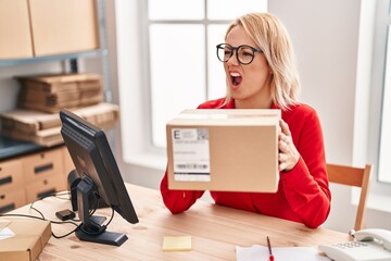 Canvas Print - Blonde woman working at small business ecommerce holding box angry and mad screaming frustrated and furious, shouting with anger. rage and aggressive concept.