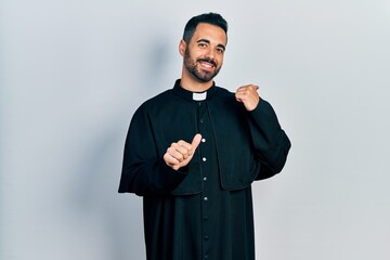 Canvas Print - Handsome hispanic man with beard wearing catholic priest robe pointing to the back behind with hand and thumbs up, smiling confident