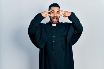 Canvas Print - Handsome hispanic man with beard wearing catholic priest robe doing peace symbol with fingers over face, smiling cheerful showing victory