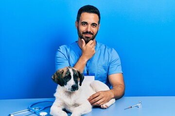 Poster - Handsome hispanic veterinary man with beard checking dog health looking confident at the camera smiling with crossed arms and hand raised on chin. thinking positive.