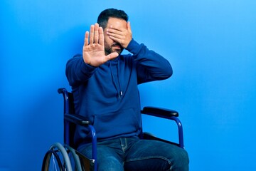 Wall Mural - Handsome hispanic man with beard sitting on wheelchair covering eyes with hands and doing stop gesture with sad and fear expression. embarrassed and negative concept.