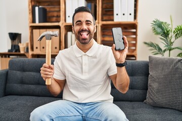 Canvas Print - Young hispanic man with beard holding hammer and broken smartphone showing cracked screen sticking tongue out happy with funny expression.