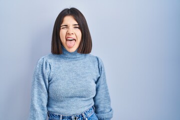 Young hispanic woman standing over blue background sticking tongue out happy with funny expression. emotion concept.