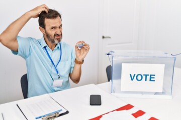 Middle age man with beard sitting by ballot holding i vote badge confuse and wondering about question. uncertain with doubt, thinking with hand on head. pensive concept.