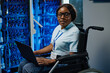 Portrait of African female IT engineer with disability sitting in wheelchair and looking at camera while working with laptop in data server room
