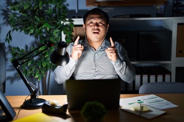 Canvas Print - Young chinese man working using computer laptop at night amazed and surprised looking up and pointing with fingers and raised arms.