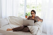 © Dmytro Hai - Positive woman in eyeglasses holding mug with coffee and using laptop on couch in living room