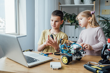 Nice interested friends smiling, chatting and working on project together using laptop. Girl helping boy in creating robot using building kit for kids on table